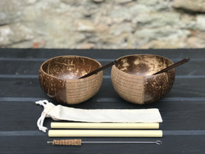 Two spoons inside two brown coconut bowls, two bamboo straws, a cleaning brush and a straw pouch on a wooden table.