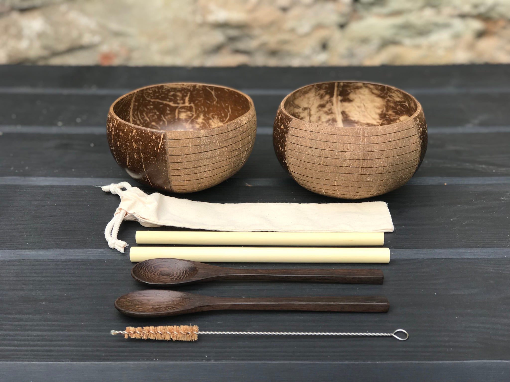 Two brown coconut bowls, two bamboo straws, two spoons, a cleaning brush and a straw pouch on a wooden table.