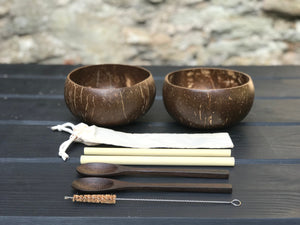 Two brown coconut bowls, two bamboo straws, two spoons, a cleaning brush and a straw pouch on a wooden table.